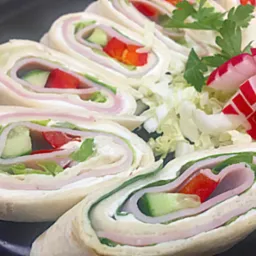 Tortilla wraps z szynką, serem, czerwoną papryką, ogórkiem i sałatą, udekorowane rzodkiewką i natką pietruszki, ułożone na czarnym talerzu.