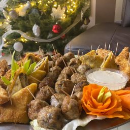 FIRA SP. Z O. O. - Taca z różnorodnymi przekąskami na przyjęcie, w tle choinka z dekoracjami, w tym roladki z farszem, mięsne kulki, smażone kawałki w panierce sezamowej, sos w przezroczystym pojemniku i dekoracja...