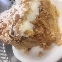 FIRA SP. Z O. O. - Kawałek szarlotki z kruszonką i bitą śmietaną, posypany cukrem pudrem na białym talerzu, widoczne jabłka w nadzieniu.