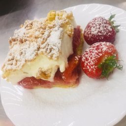 FIRA SP. Z O. O. - Kawałek ciasta rabarbarowego pod kruszonką, przekładany kremem, posypany cukrem pudrem, obok leżą dwie świeże truskawki również oprószone cukrem pudrem, na białym talerzu.