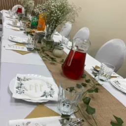 Uroczysty stół nakryty białym obrusem z bieżnikiem jutowym, dekorowany zielonymi pnączami, wazonami z gipsówką, szklankami, dzbankiem z czerwonym napojem i serwetkami z napisem 'Pierwsza Komunia...