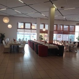 FIRA SP. Z O. O. - Sala bankietowa z długimi stołami nakrytymi białymi obrusami, krzesła w białych pokrowcach, czerwone i czarne fotele ustawione w rzędach, okrągłe papierowe lampiony podwieszone do sufitu, duże okna...