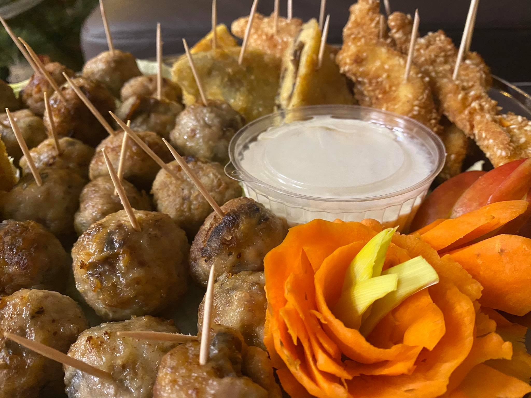 Dekoracyjnie ułożone przekąski na tacy: mini klopsiki z wykałaczkami, smażone kieszonki, paluszki w sezamie, dip w plastikowym pojemniku oraz róża z marchewki.