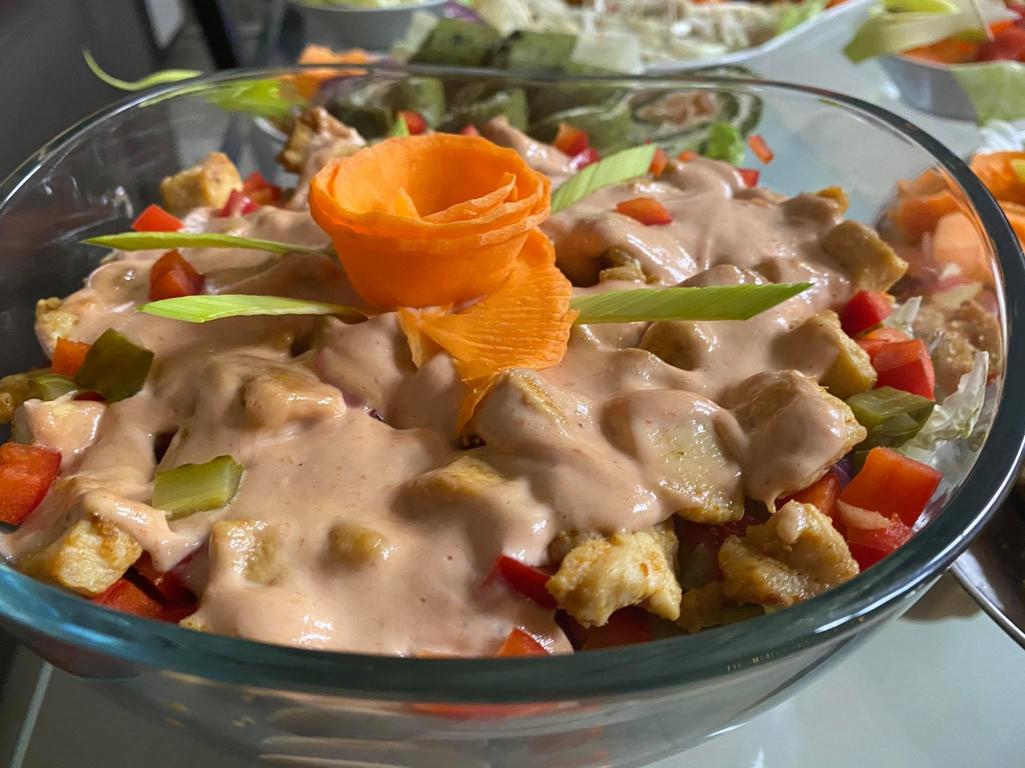 Sałatka warstwowa w szklanej misce, udekorowana kwiatem z marchewki i polana sosem, widoczne kawałki papryki i ogórka.