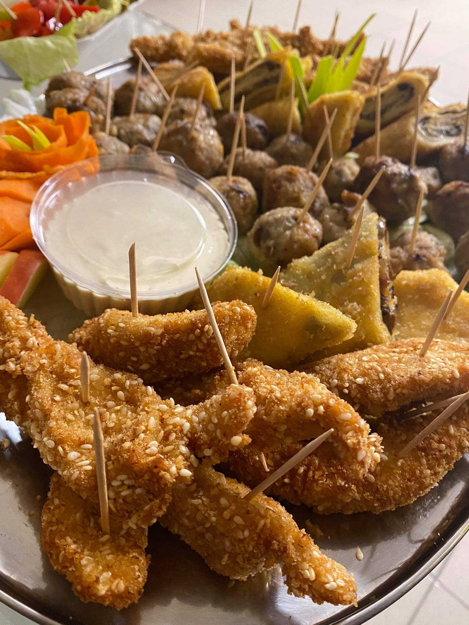 Srebrna patera z różnorodnymi przekąskami na jeden kęs, w tym nuggetsy w sezamie na wykałaczkach, pieczarki, krokiety i sos, udekorowana rzeźbą z marchewki.