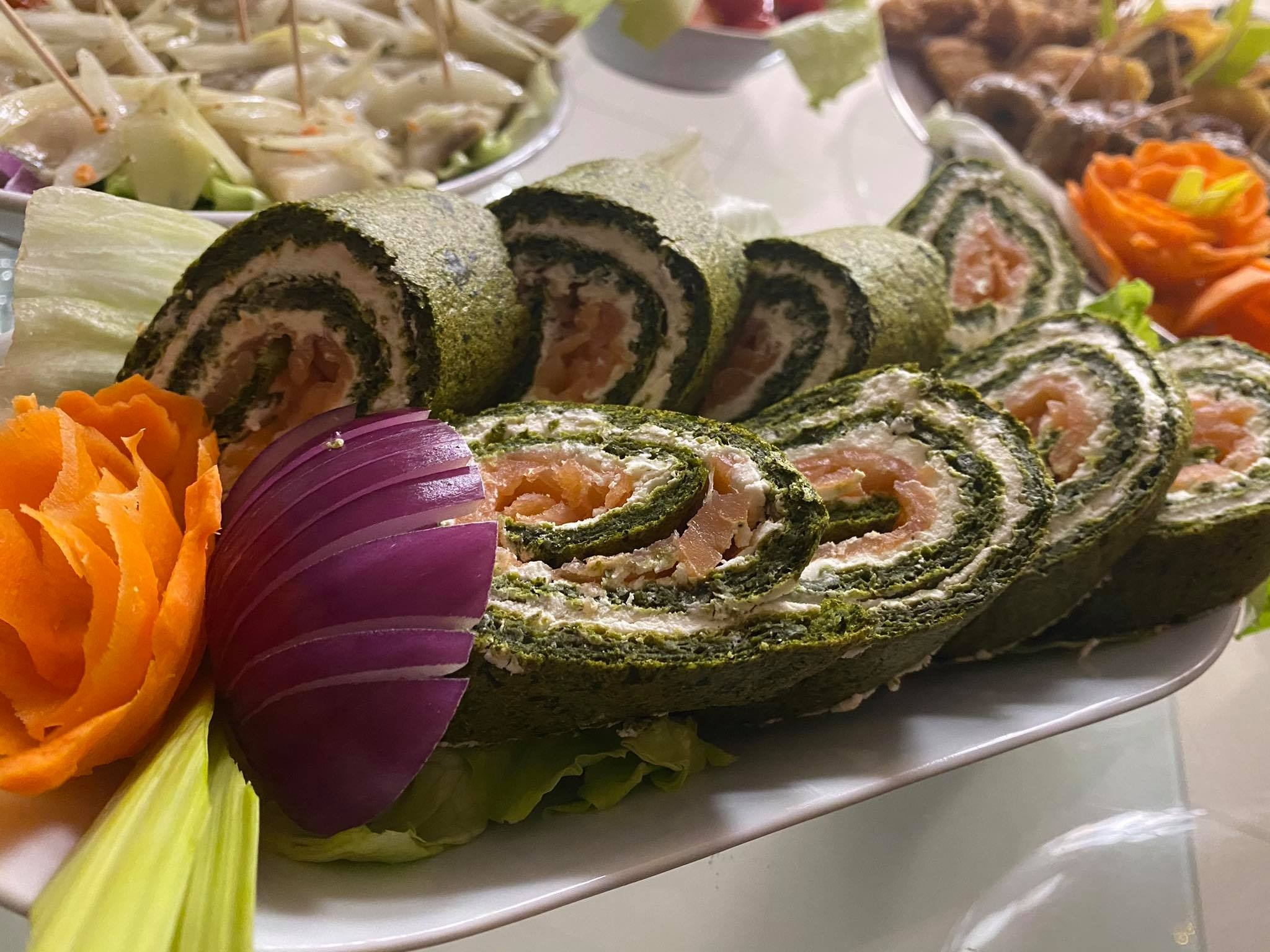 Dekoracyjnie ułożone roladki ze szpinakowego ciasta z nadzieniem serowym i łososiem, ozdobione rzeźbioną marchewką i czerwoną cebulą na liściach sałaty.