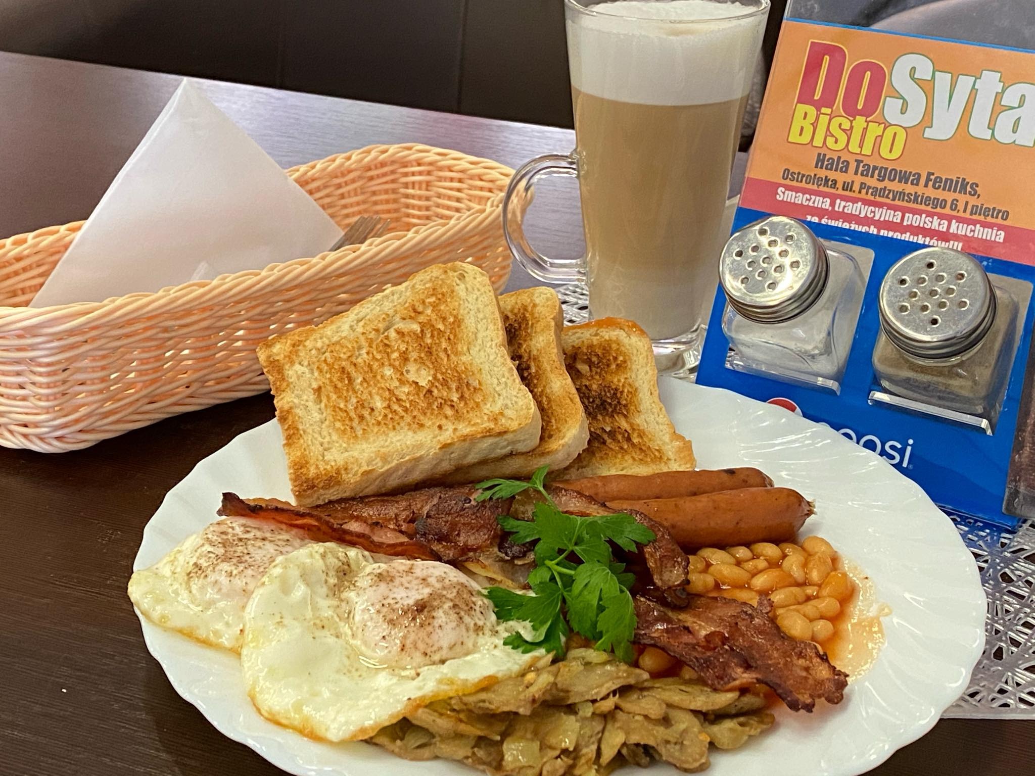 Pełny angielski śniadanie na białym talerzu: tosty, jajka sadzone, bekon, kiełbaski, fasolka i smażone pieczarki, uzupełnione latte w szklance i serwetkami w koszyczku.