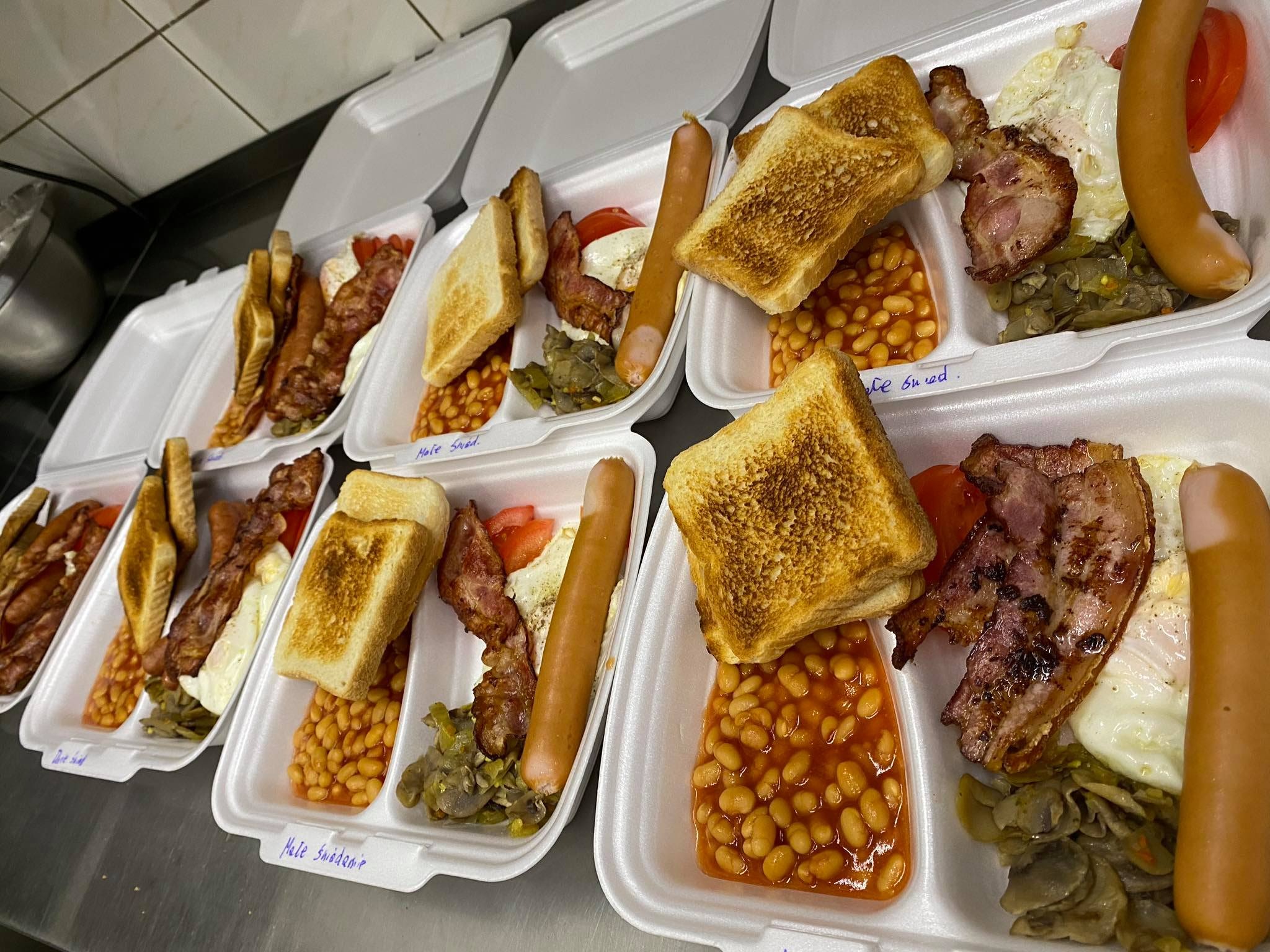 Rzędy styropianowych pudełek z pełnym angielskim śniadaniem: fasolka, bekon, jajko sadzone, kiełbasa, pomidor i grzanka, przygotowane do transportu.