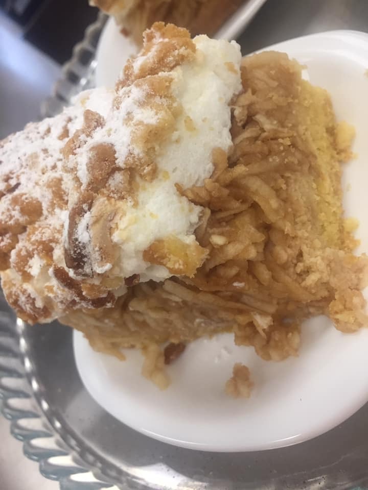 Kawałek szarlotki z kruszonką i bitą śmietaną, posypany cukrem pudrem na białym talerzu, widoczne jabłka w nadzieniu.