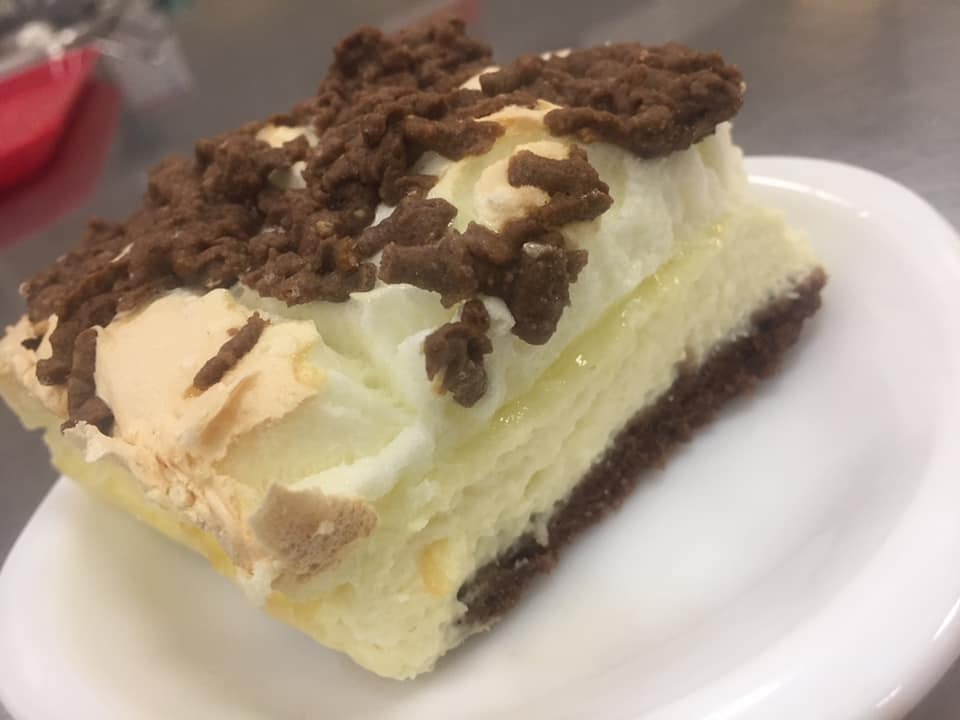 Kawałek ciasta z ciemnym spodem, kremowym środkiem i posypką z czekoladowych wiórków na białym talerzu.