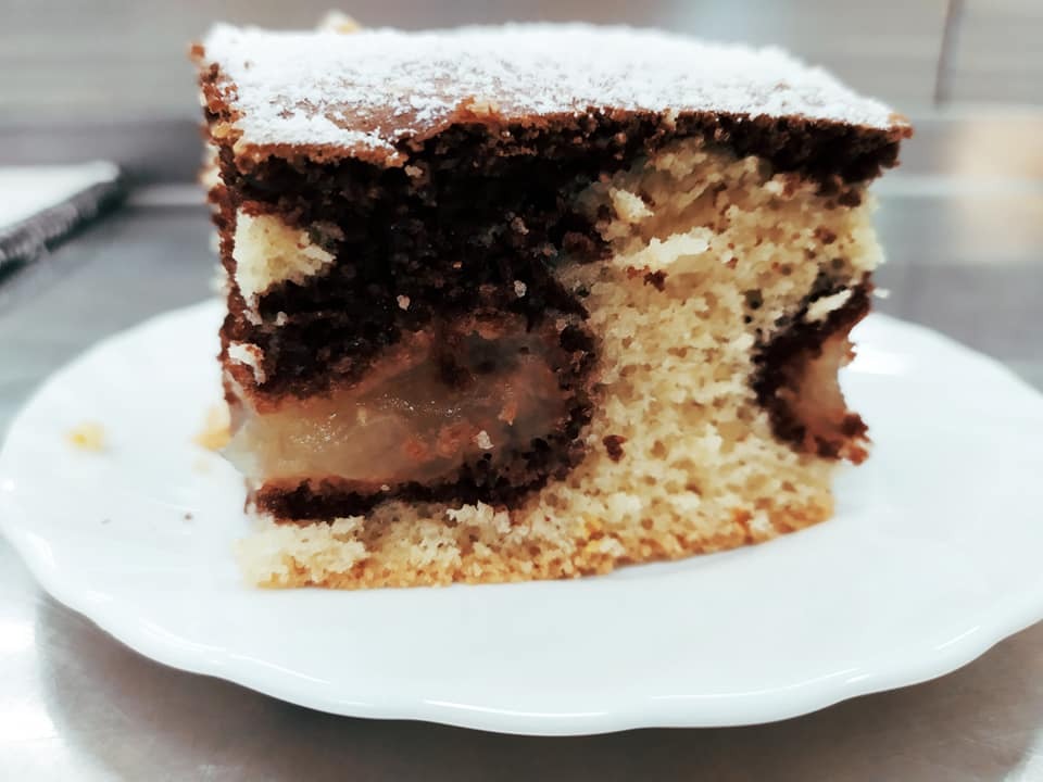 Kawałek dwukolorowego ciasta z jabłkami i cukrem pudrem na białym talerzu.