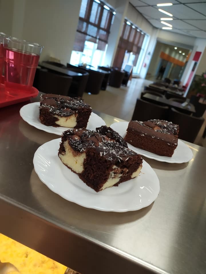 Trzy kawałki brownie z kremem i posypką kokosową na białych talerzykach, ustawione na metalowym blacie w przestronnym pomieszczeniu z widocznymi stolikami i oknami.