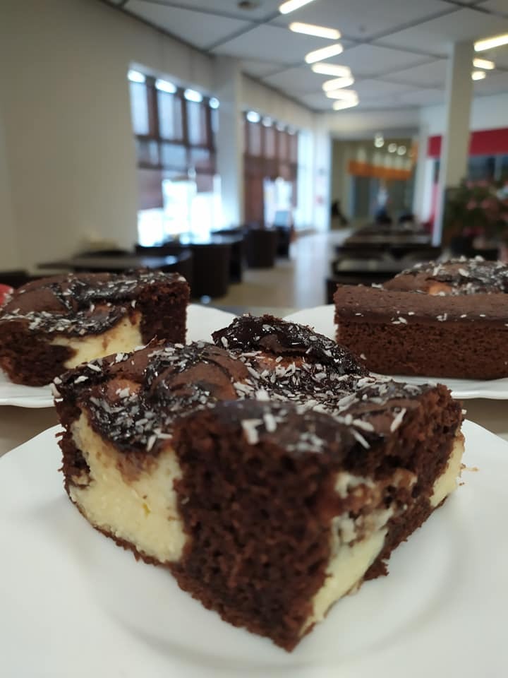 Kawałki ciasta brownie z kremem serowym i posypką kokosową na białych talerzach w jasnym wnętrzu biurowym.