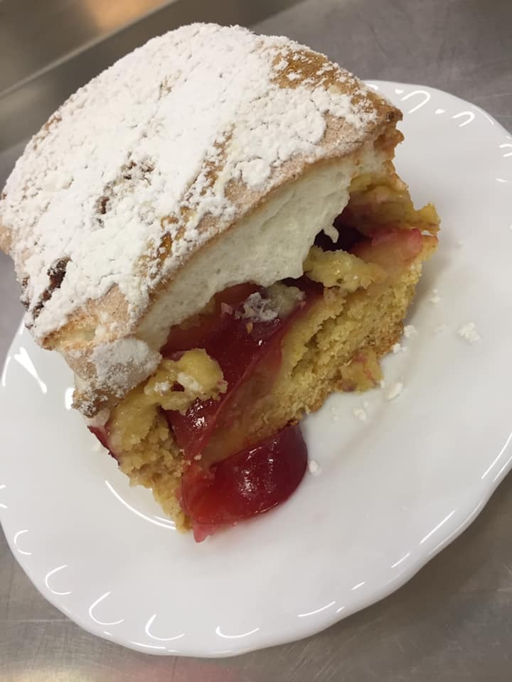 Kawałek domowego ciasta ze śliwkami i bitą śmietaną posypany cukrem pudrem, ułożony na białym, falowanym talerzu.