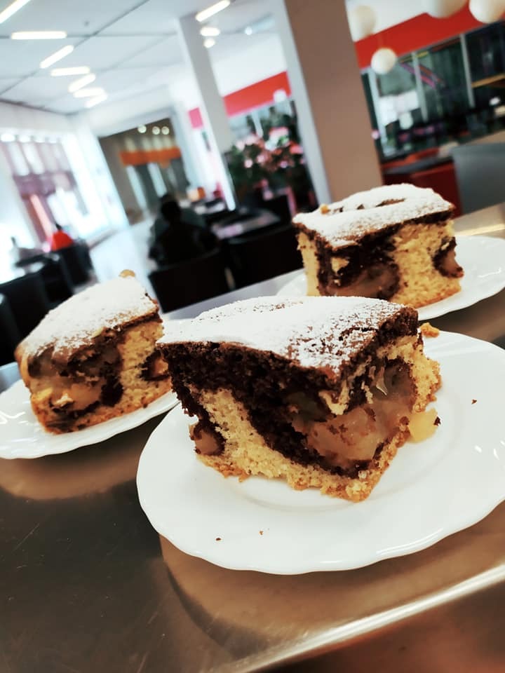 Trzy porcje marmurkowego ciasta jabłkowego posypane cukrem pudrem na białych talerzykach, w tle jasne biuro z siedzącymi osobami.