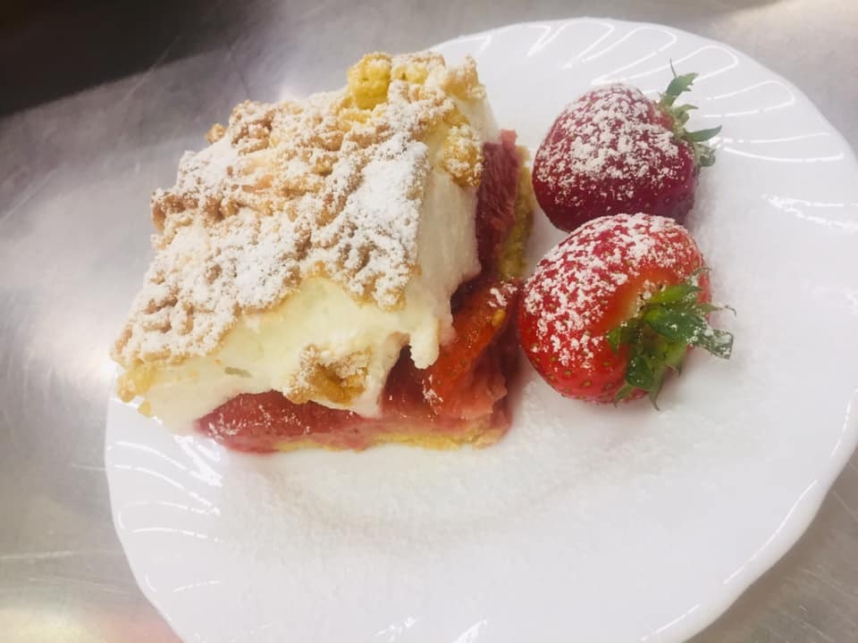 Kawałek ciasta rabarbarowego pod kruszonką, przekładany kremem, posypany cukrem pudrem, obok leżą dwie świeże truskawki również oprószone cukrem pudrem, na białym talerzu.