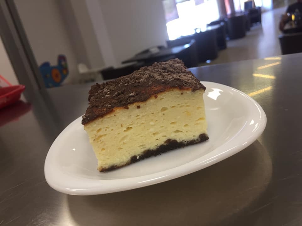 Kwadratowy kawałek sernika z kruszonką czekoladową na białym talerzu, widoczny na metalowym blacie stołu, w tle rozmyte krzesła i okno.