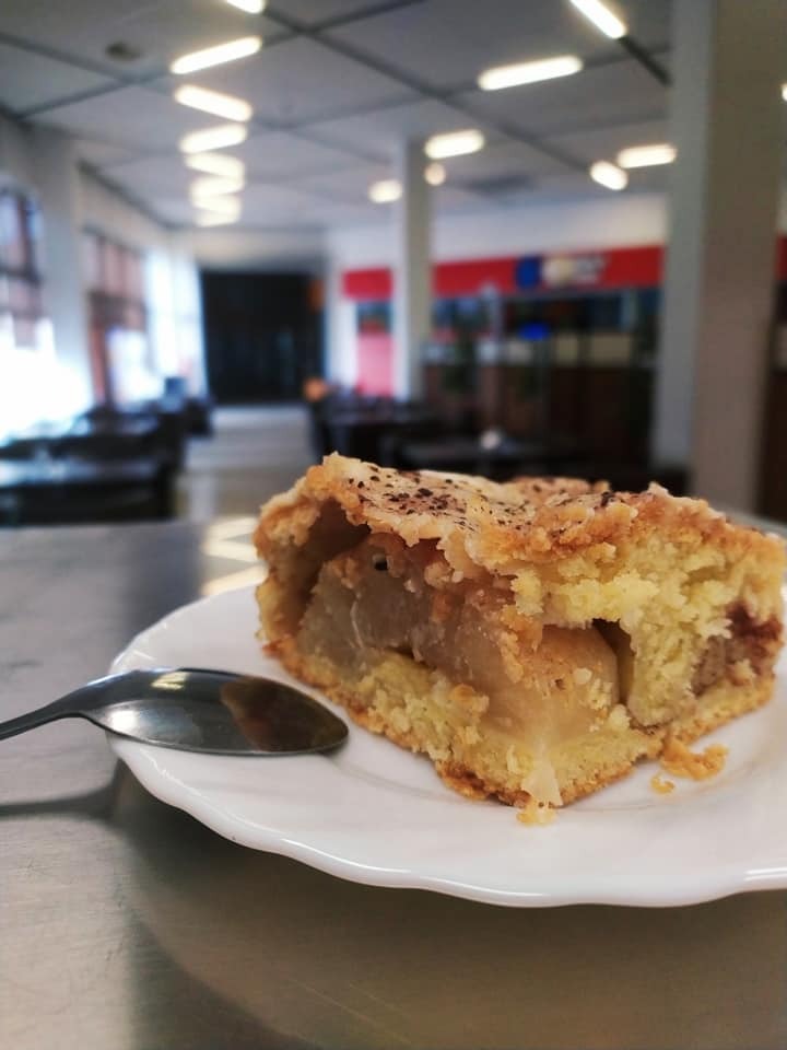 Kawałek szarlotki z kruszonką, posypany cynamonem, podany na białym talerzu obok łyżeczki, w tle wnętrze kawiarni.