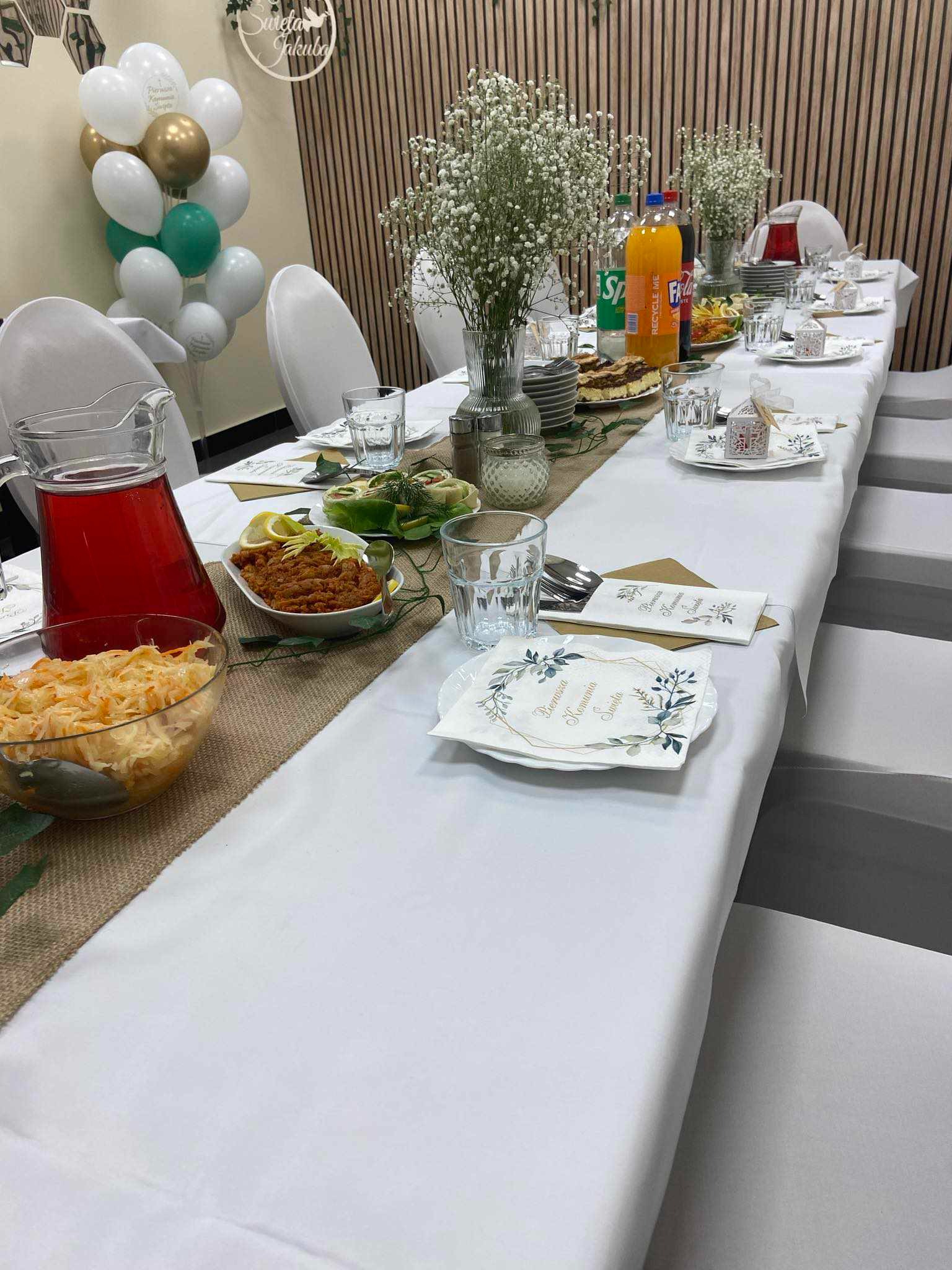 Długi stół nakryty białym obrusem i jutowym bieżnikiem, udekorowany kwiatami gipsówki, potrawami w misach i dzbanku z czerwonym napojem, z balonami w tle.