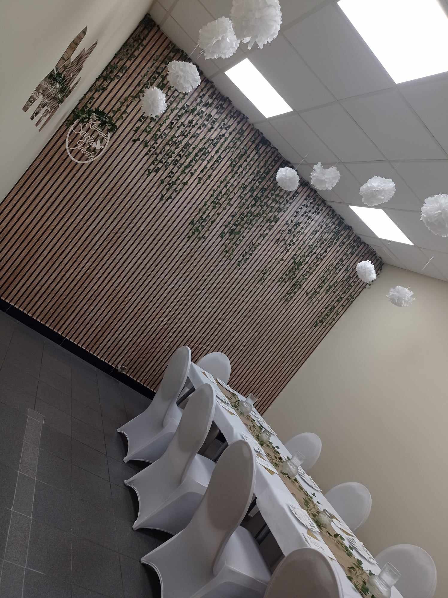 Sala bankietowa z dekoracją na Komunię Świętą: drewniana ściana z zielonymi pnączami i białymi pomponami, stół z białym obrusem i jutowym bieżnikiem, białe krzesła z pokrowcami.