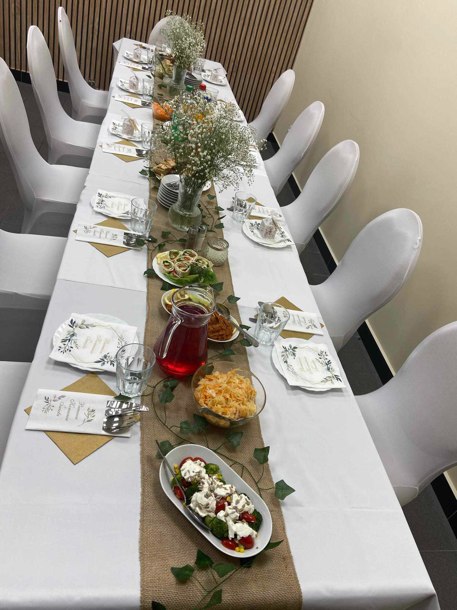 Długi stół nakryty białym obrusem z jutowym bieżnikiem, udekorowany kompozycjami kwiatowymi z gipsówki, zastawą, serwetkami z nadrukiem, karafką z czerwonym napojem i różnorodnymi potrawami...