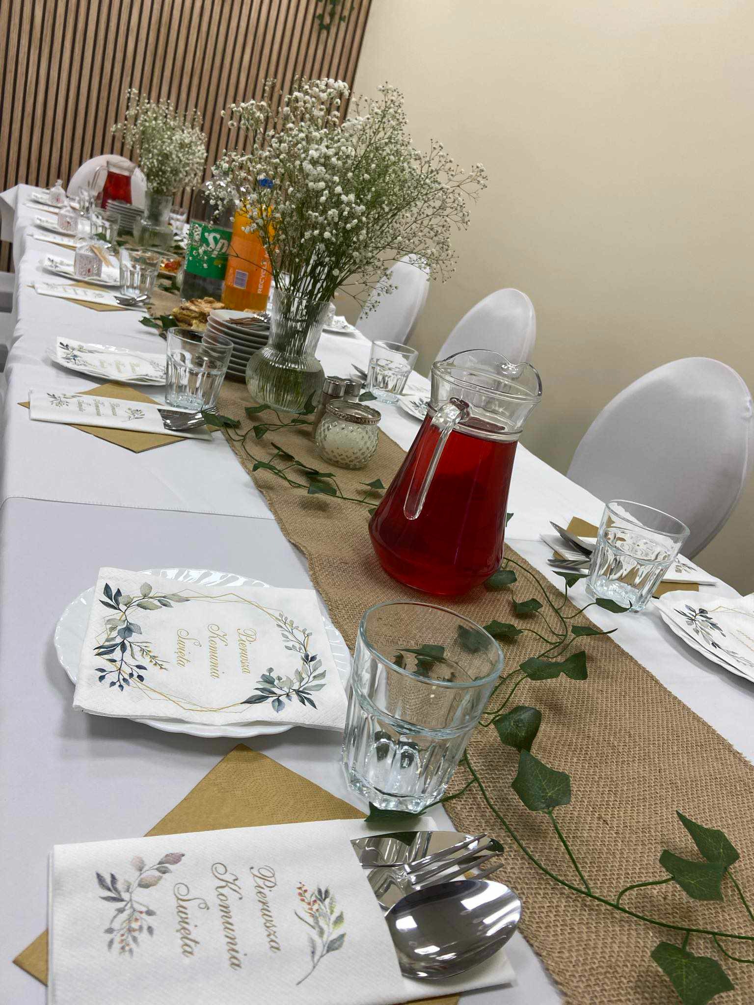 Uroczysty stół nakryty białym obrusem z bieżnikiem jutowym, dekorowany zielonymi pnączami, wazonami z gipsówką, szklankami, dzbankiem z czerwonym napojem i serwetkami z napisem 'Pierwsza Komunia...