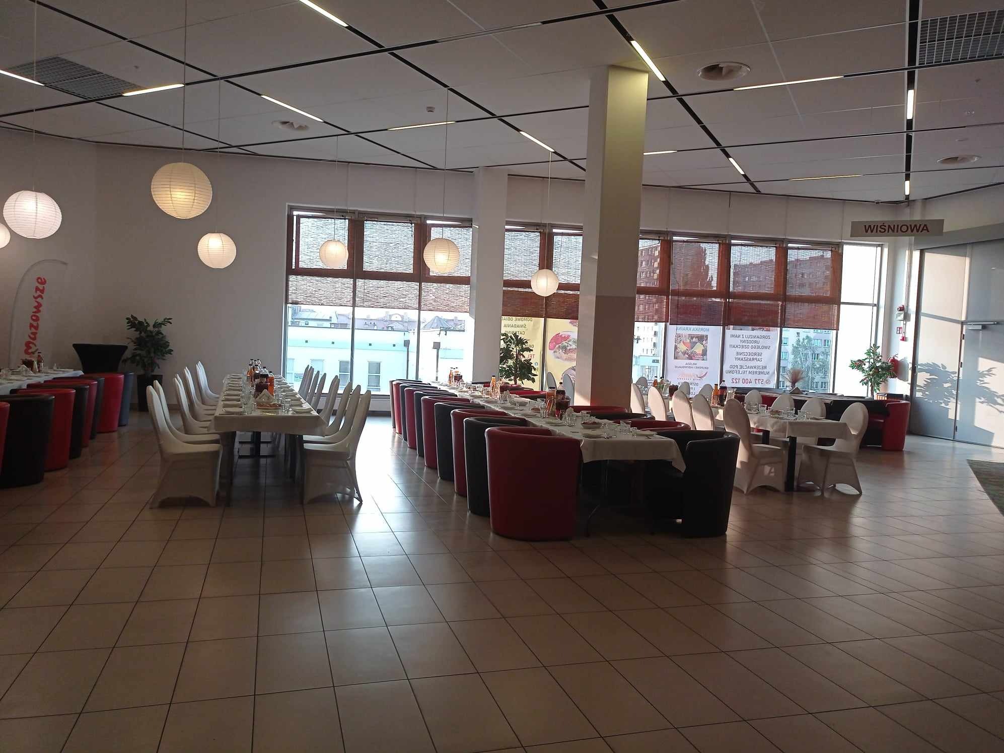 Sala bankietowa z długimi stołami nakrytymi białymi obrusami, krzesła w białych pokrowcach, czerwone i czarne fotele ustawione w rzędach, okrągłe papierowe lampiony podwieszone do sufitu, duże okna...