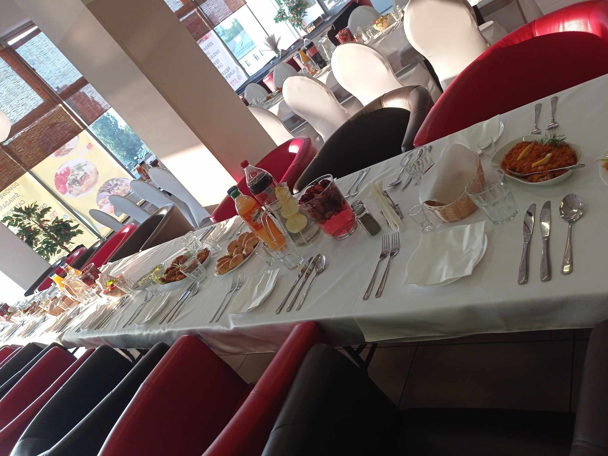 Długi stół z białym obrusem, nakryty zastawą, napojami i przekąskami, przygotowany dla gości; widoczne krzesła w kolorach czerwonym i czarnym.