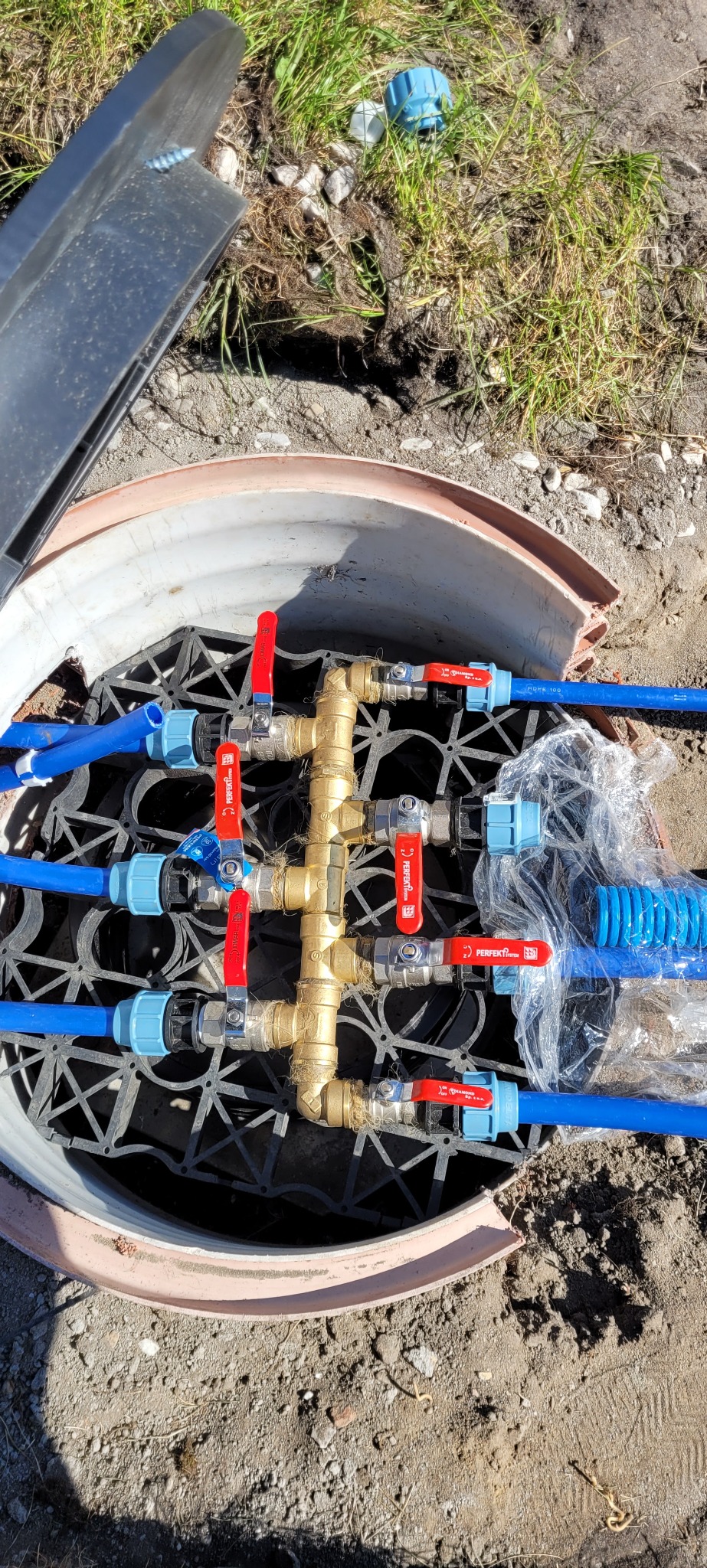 Węzeł zaworowy systemu nawadniania w studzience z niebieskimi rurami i zaworami z czerwonymi rączkami, widoczne elementy montażowe i opakowanie ochronne, z otwartą pokrywą studzienki