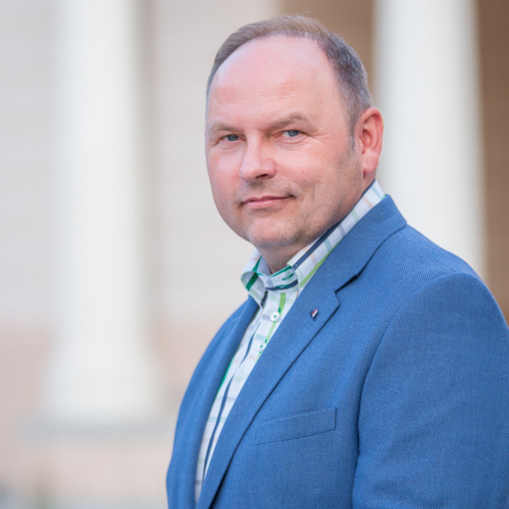 Portret mężczyzny w niebieskiej marynarce i wzorzystej koszuli, stojącego na tle kolumn.