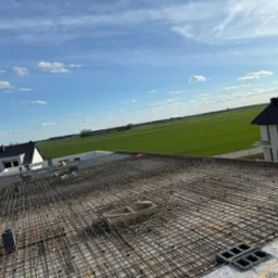 Zbrojenie stropu budynku w trakcie budowy, widok z góry na siatkę z prętów stalowych na tle zielonego pola i błękitnego nieba z chmurami.