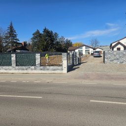 BARTOSZ ZDUNEK USŁUGI OGÓLNO-BUDOWLANE - Pracownik w jaskrawej kamizelce wyrównuje teren podłoża przed posesją z nowym, szarym ogrodzeniem z elementami imitującymi kamień i siatką, widok z ulicy.