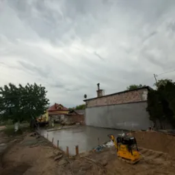 Wylany fundament pod budowę z żółtą zagęszczarką obok. Widoczny szalunek, budynek w tle i pochmurne niebo.