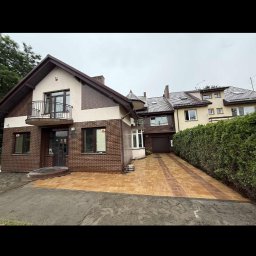 BARTOSZ ZDUNEK USŁUGI OGÓLNO-BUDOWLANE - Nowa, brązowo-żółta kostka brukowa przed domem z cegły. Ułożona wzorem w pasy, prowadzi do garażu. Widoczne ślady ziemi po bokach. Dom dwukondygnacyjny z balkonem.