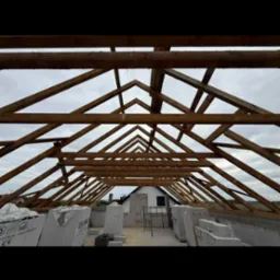 Konstrukcja więźby dachowej z drewnianych belek na budowie domu, widok z wnętrza. Ułożone bloczki i materiały budowlane w tle. Niebo widoczne przez konstrukcję.