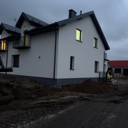 Bartosz Zdunek - Biały dom z ciemną dachówką i wykończeniem, widoczny fragment wykopu i sprzętu budowlanego, pochmurne niebo w tle. Świecące okna.