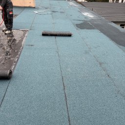 BARTOSZ ZDUNEK USŁUGI OGÓLNO-BUDOWLANE - Prace na dachu płaskim: fragment pokrycia z papy termozgrzewalnej w kolorze niebiesko-szarym, częściowo ułożonej, z widocznym pracownikiem i rolką materiału.