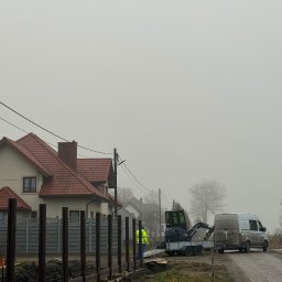 BARTOSZ ZDUNEK USŁUGI OGÓLNO-BUDOWLANE - Montaż ogrodzenia panelowego z drewnianymi słupkami i podmurówką. W tle dom jednorodzinny i mini koparka na przyczepie. Prace ziemne w toku, pochmurny dzień.