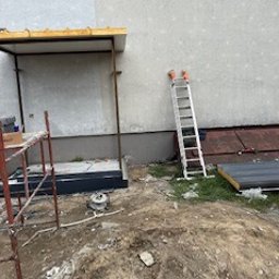 BARTOSZ ZDUNEK USŁUGI OGÓLNO-BUDOWLANE - Budowa ganku przy ścianie budynku z drabina i rusztowaniem. Widoczny fragment ocieplenia. Prace w toku, ziemia wokół. Ujęcie z dołu, lekko z prawej strony.