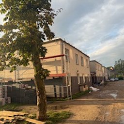 BARTOSZ ZDUNEK USŁUGI OGÓLNO-BUDOWLANE - Budynek w trakcie prac elewacyjnych z rusztowaniem, widok z dołu, z boku drzewo, w tle pochmurne niebo. Częściowo ocieplona ściana, widoczne materiały budowlane.