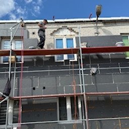 BARTOSZ ZDUNEK USŁUGI OGÓLNO-BUDOWLANE - Praca na rusztowaniu: ocieplanie elewacji budynku styropianem grafitowym. Widoczne okna, fragment dachu i niebo. Jeden pracownik na górze, drugi na dole.