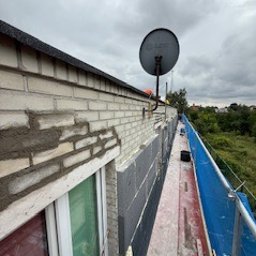 BARTOSZ ZDUNEK USŁUGI OGÓLNO-BUDOWLANE - Ściana budynku w trakcie ocieplania: widoczne fragmenty muru, warstwa izolacji termicznej i rusztowanie z niebieską siatką ochronną. Antena satelitarna na ścianie.