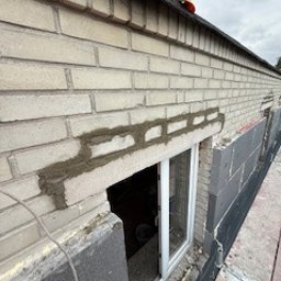 Bartosz Zdunek - Fragment ściany z jasnej cegły z oknem, częściowo ocieplonej szarym styropianem. Na górze żółty kask budowlany. Widoczne ślady prac murarskich.