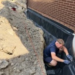 BARTOSZ ZDUNEK USŁUGI OGÓLNO-BUDOWLANE - Pracownik w wykopie układa folię kubełkową przy fundamencie ceglanego budynku. Widoczne zabezpieczenie przeciwwilgociowe i drenaż. Prace ziemne w toku.