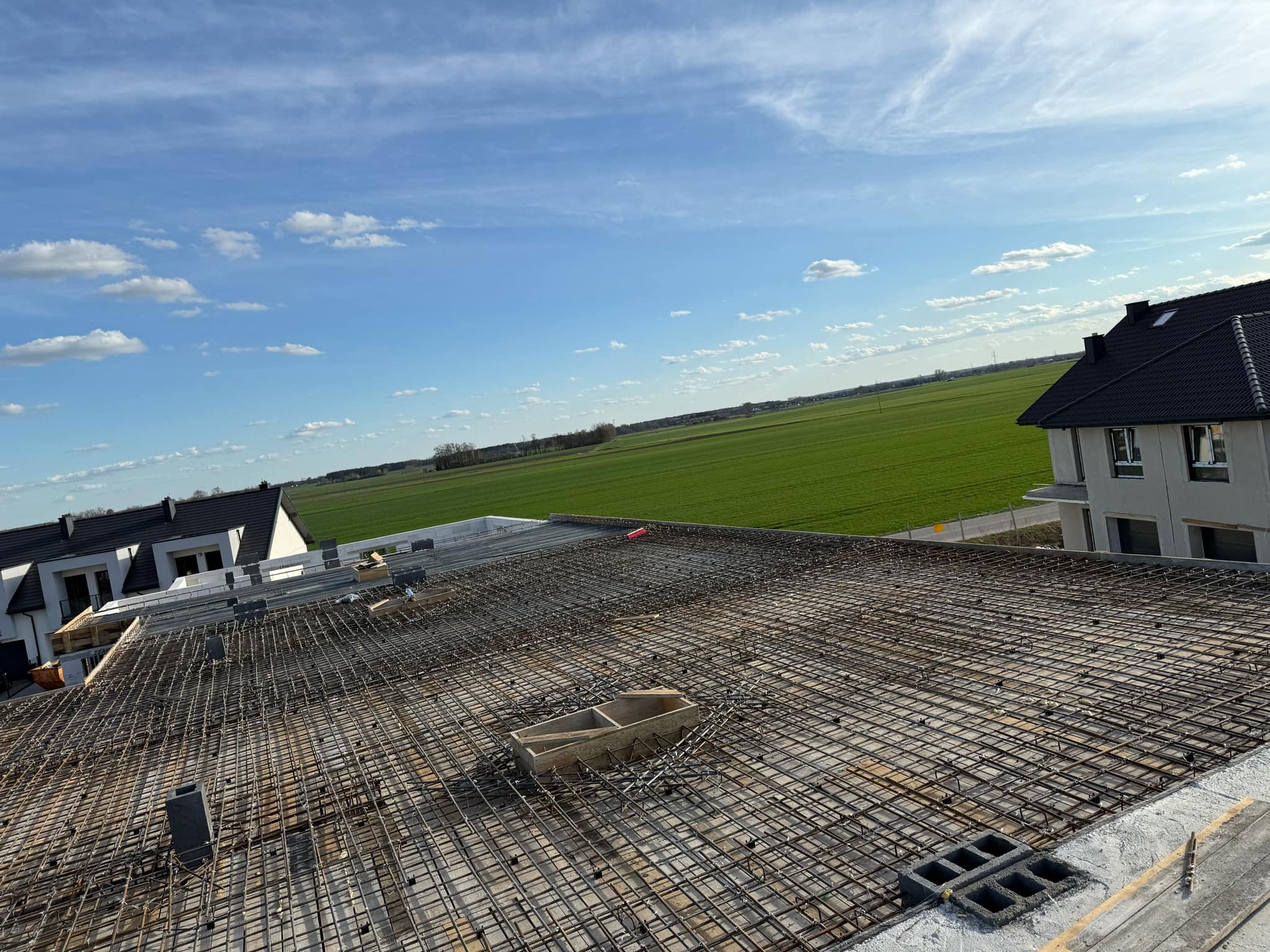 Zbrojenie stropu budynku w trakcie budowy, widok z góry na siatkę z prętów stalowych na tle zielonego pola i błękitnego nieba z chmurami.