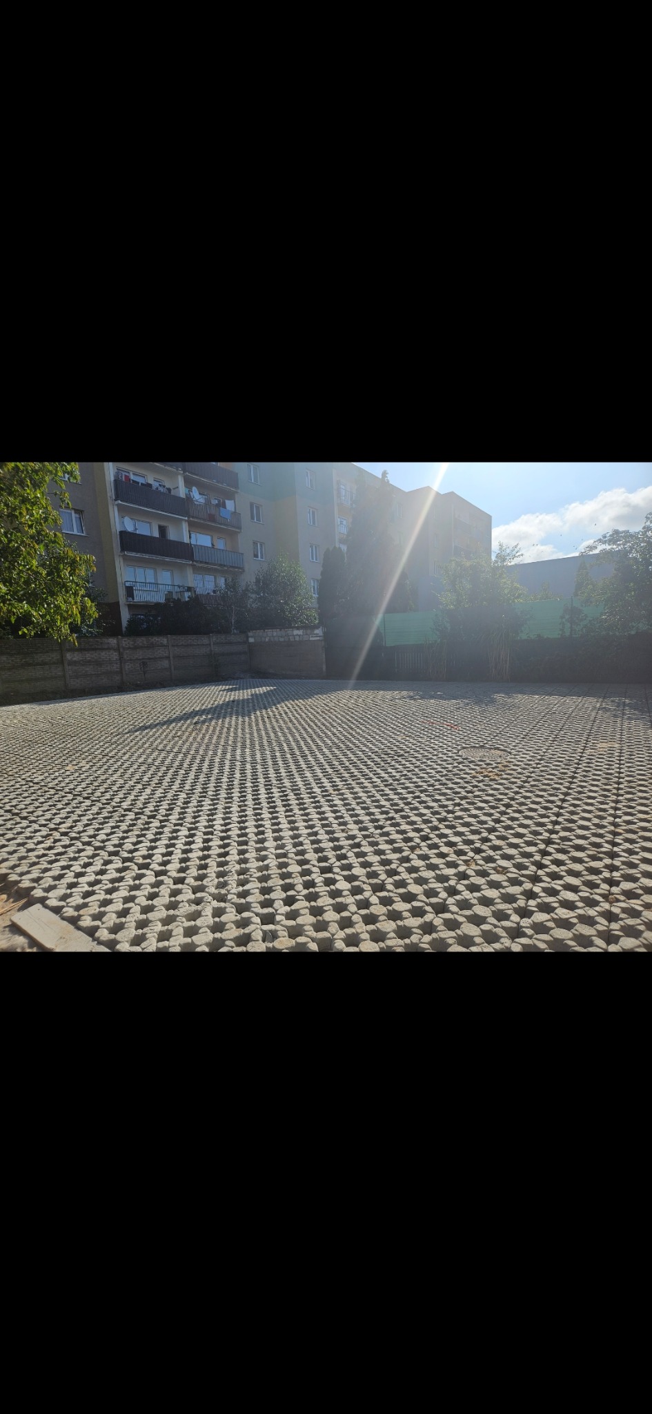 Ułożona nawierzchnia z ażurowych betonowych płyt na parkingu przed budynkiem mieszkalnym, widoczne drzewa i fragment nieba.
