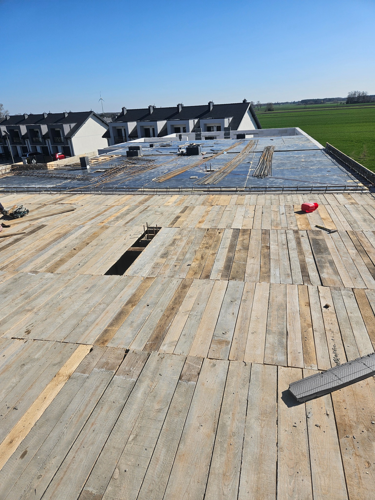 Wysoki kąt widzenia na drewniany szalunek stropu budynku w trakcie budowy, z widocznymi prętami zbrojeniowymi i elementami konstrukcyjnymi, w tle zabudowa szeregowa i pola uprawne.