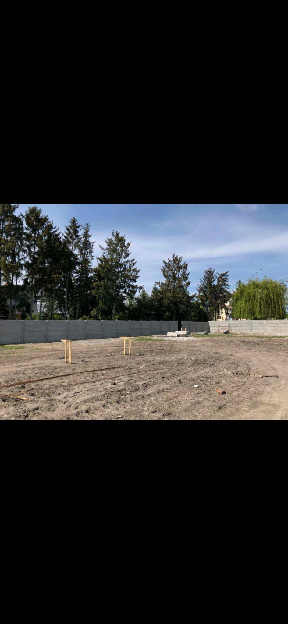 Przygotowany teren pod budowę z widocznymi drewnianymi słupkami i betonowym ogrodzeniem w tle.