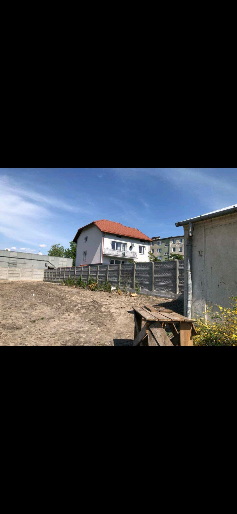 Działka z wyrównanym terenem przygotowana pod budowę, ogrodzona betonowym płotem z ozdobnymi panelami, w tle dom jednorodzinny z balkonem i budynek wielorodzinny.