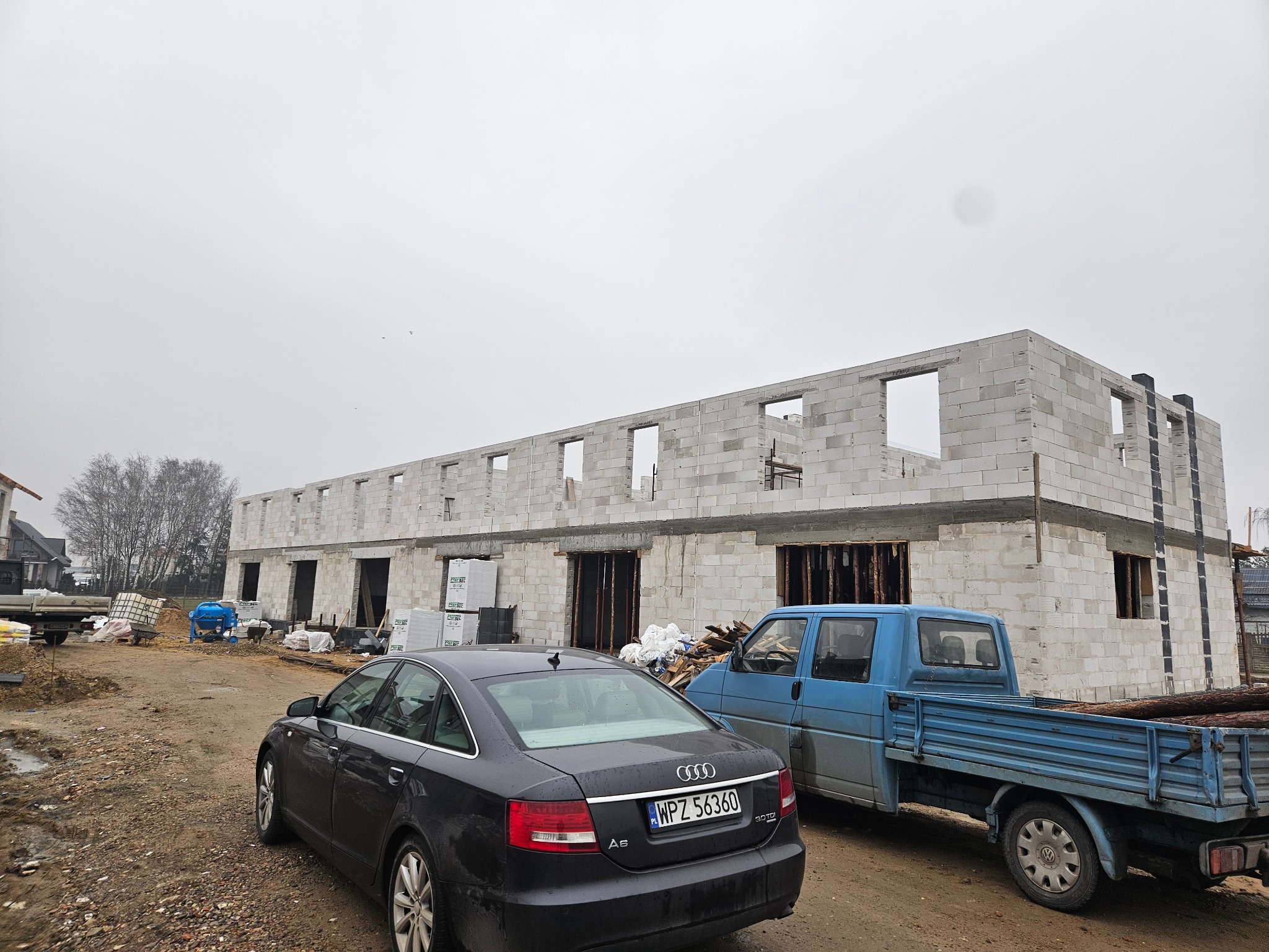 Wznoszenie dwukondygnacyjnego budynku z betonu komórkowego, widoczne szare ściany w stanie surowym, otwory okienne i drzwiowe, na pierwszym planie zaparkowane samochody: czarne Audi A6 i niebieski...